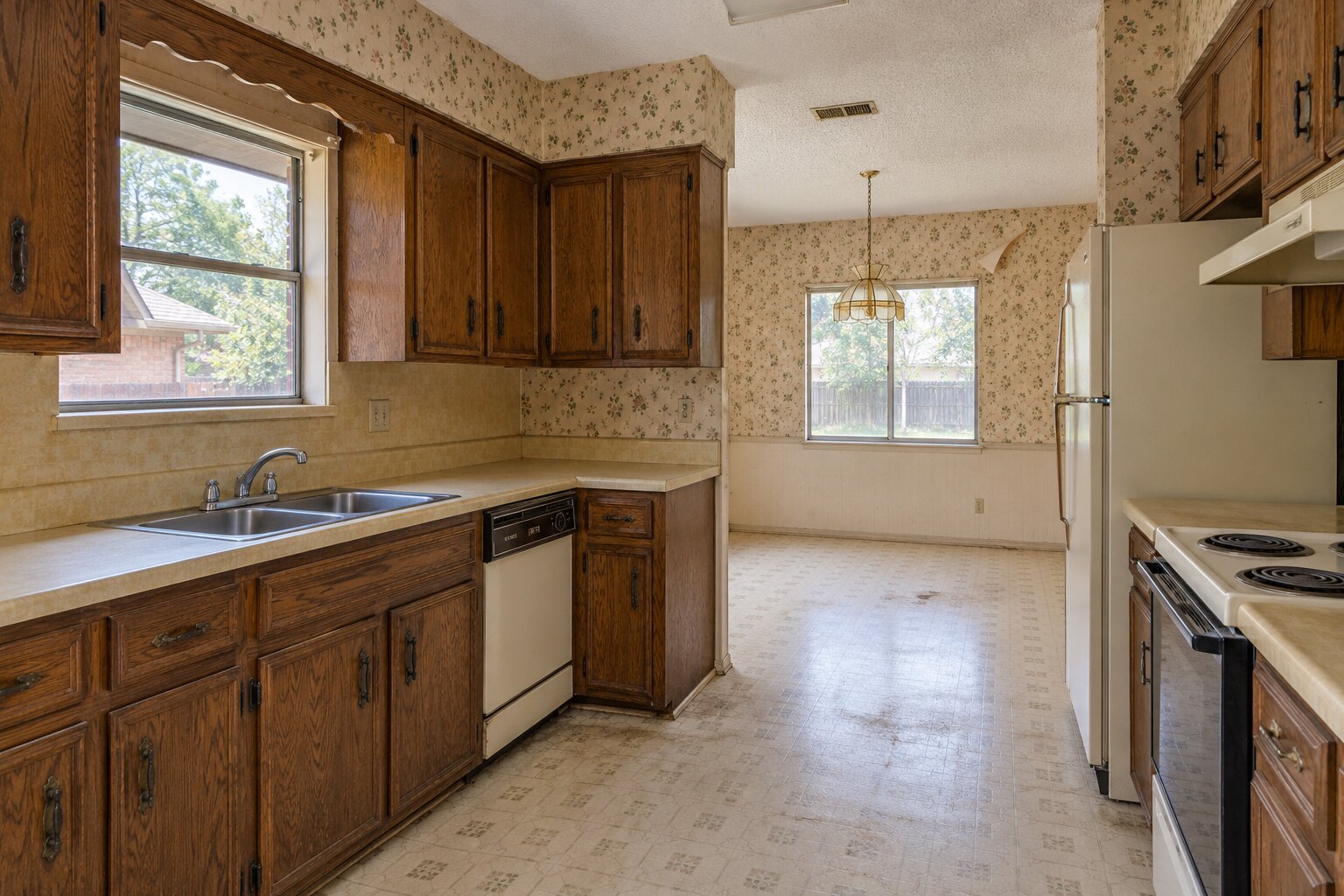 An unrenovated kitchen with original cabinets, dated wallpaper, and worn flooring